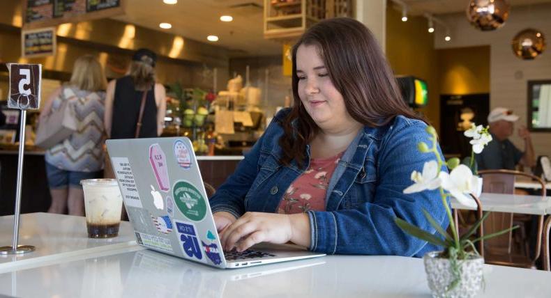 A student types on their laptop and sits at a table in a cafe.